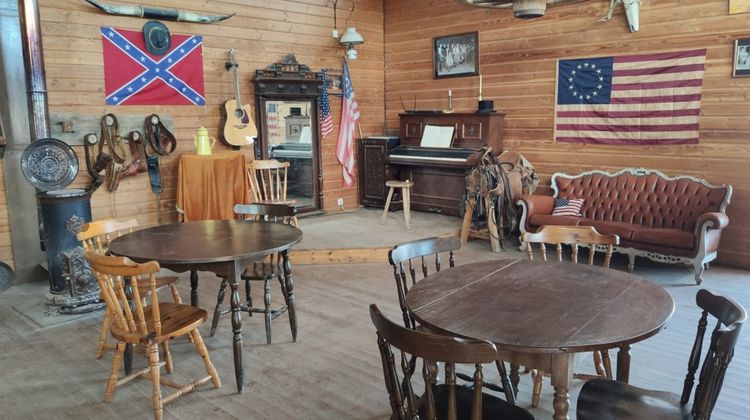 Vue d'un côté du saloon, avec tables, chaises, piano, drapeau des confédérés et à 13 étoiles, vieux poêle, etc.